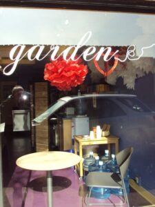 A storefront window with "garden" written in cursive. Inside, water dispensers, a small wooden table with items, and red and white paper decorations are visible. A car is parked outside.