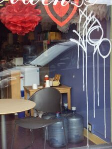 A small café interior with a table, chairs, water cooler bottles, and a counter in the background. Seen through a glass window with white graffiti and red paper decorations hanging from the ceiling.