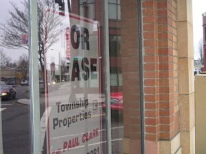 A storefront with a "FOR LEASE Township Properties Call PAUL CLARK" sign in the window, reflecting the street and nearby buildings.