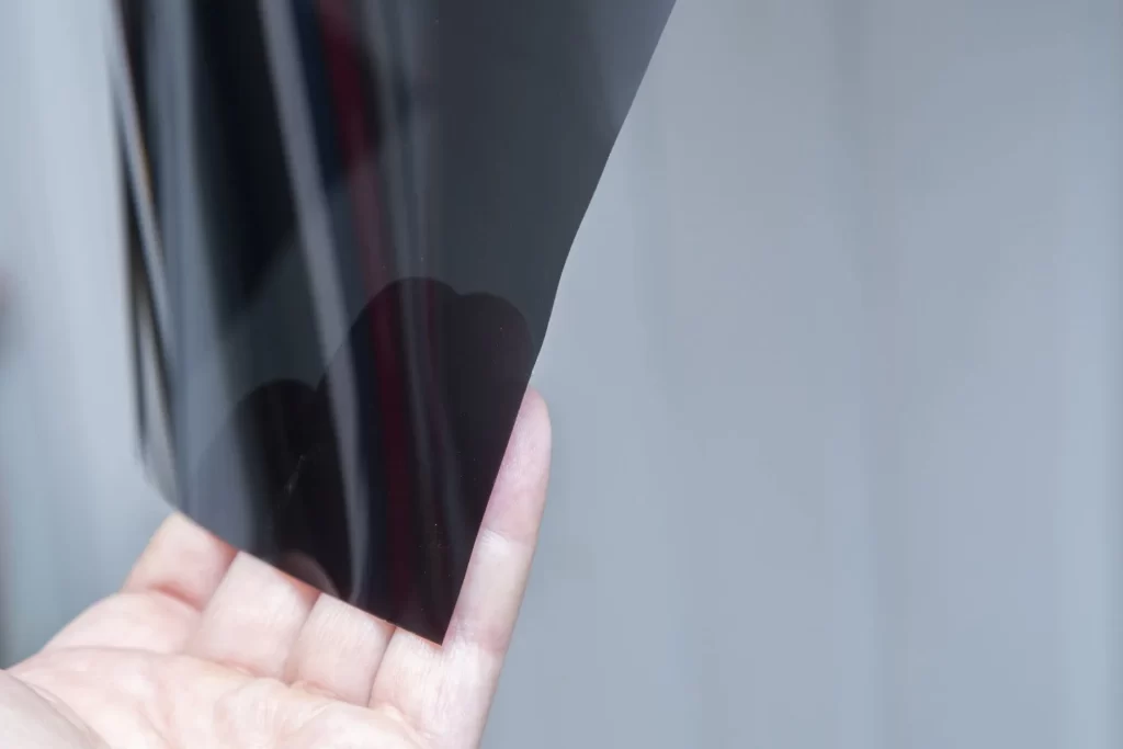 A person holds a transparent sheet of dark material between their fingers against a blurry grey background.