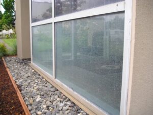 A large window with cracked glass, bordered by a smooth frame, and set next to a landscaping of rocks and mulch.