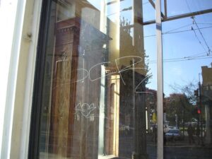 A glass window displays graffiti in white writing. The reflection shows an urban street scene with parked cars, street signs, and trees.